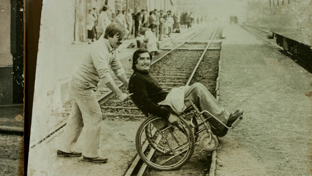 Franco Malerba, in una foto in bianco e nero, che attraversa i binari della ferrovia in carrozzina spinto da un'altra persona